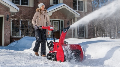 Wide view of a woman clearing the driveway with a snowblower on a sunny day. Behind her is a nice, upscale red brick house.  