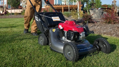 Un employé en entretien paysager pousse une tondeuse Honda près d’un buisson sur un terrain de golf. Un autre employé et un golfeur se trouvent en arrière-plan.
