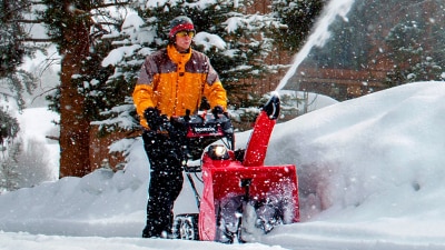 Plan étendu d’un homme qui déblaie une entrée avec une souffleuse à neige juste à l’extérieur d’un chalet.