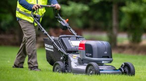 Vue de trois-quart avant droite d'un ouvrier poussant une tondeuse à gazon à baterie sur la pelouse. 