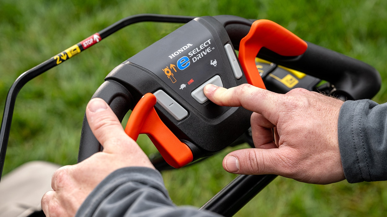 Close up of a person pushing the e-select drive button located on the lawn mower's handle.